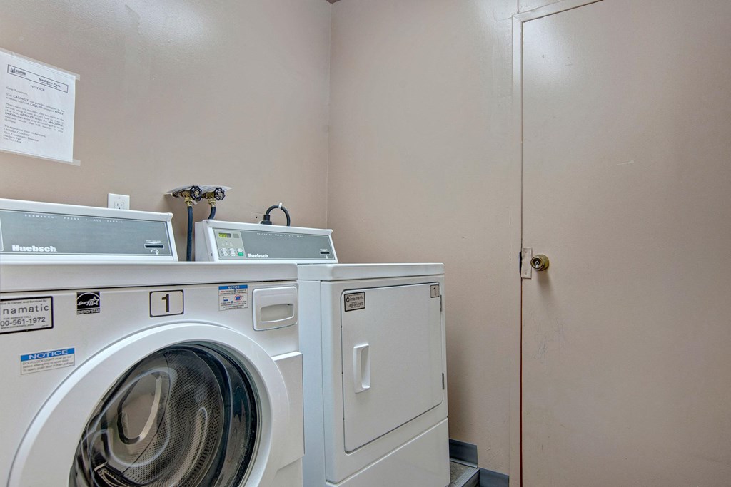 Laundry room here at Madison Park Apartment Homes with a white front-loading washing machine and dryer against a beige wall. Notice sign on the wall adds a utilitarian feel.