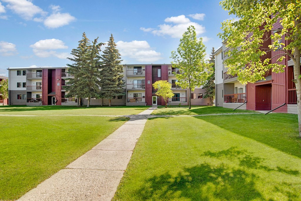 Madison Park Apartment Homes buildings with red accents overlook a lush, green courtyard on a sunny day. Tall trees and a clear path create an inviting atmosphere.