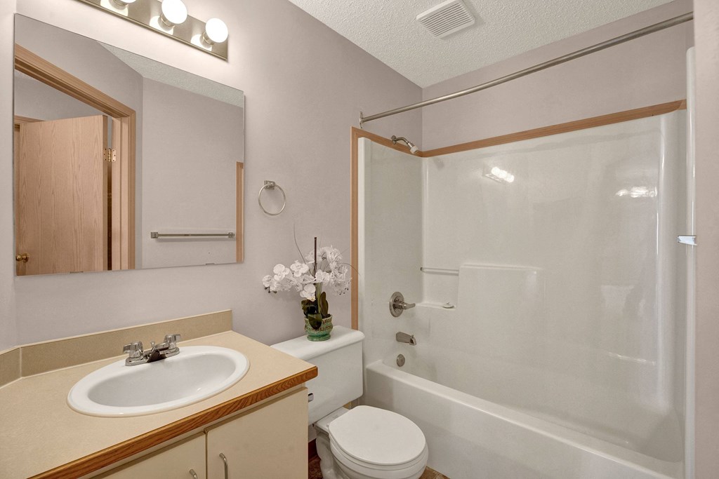 A clean, minimalist bathroom here at Manning Crossing Apartment Homes with a beige counter, white sink, and mirror. A white orchid decorates next to a toilet. A bathtub with a shower curtain rail is in the corner.