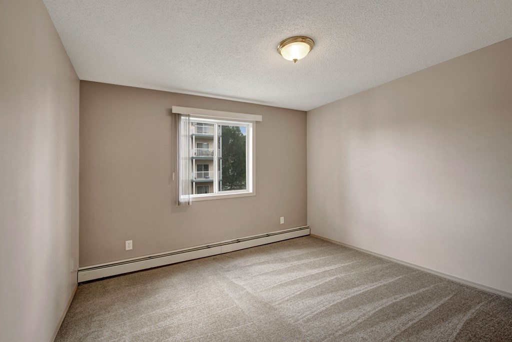 Empty room here at Manning Crossing Apartment Homes with beige walls and carpet, featuring a large window with blinds, allowing natural light. The space feels clean and minimal.