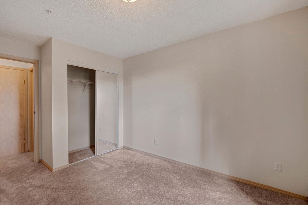 Empty bedroom here at Manning Crossing Apartment Homes with beige carpet and pale walls, mirror sliding closet doors, and a wooden door leading to another room. Calm and neutral atmosphere.