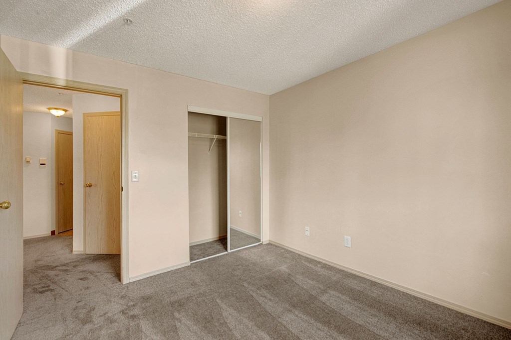 Empty room here at Manning Crossing Apartment Homes with beige walls and gray carpet, featuring mirrored closet doors on the right and a slightly ajar wooden door to the left. Warm, neutral tone.