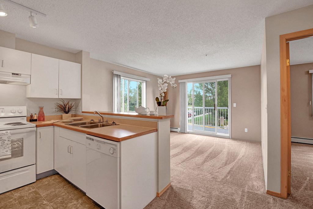 Bright, modern kitchen and living room here at Manning Crossing Apartment Homes with white cabinets, beige countertops, carpeted floor, large windows, and sliding glass door to a balcony.