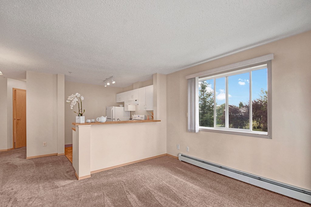 Cozy apartment interior here at Manning Crossing Apartment Homes with beige walls and carpeted floor. A small kitchen area features white cabinets and a breakfast bar with an orchid plant. Large window, bright natural light.