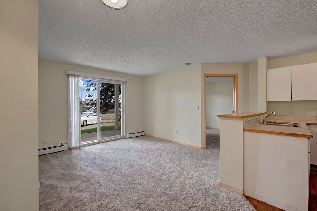 Spacious, empty living room here at Manning Crossing Apartment Homes with light gray carpeting, beige walls, and sliding glass door leading to a patio. Adjacent open kitchen and doorway to another room.