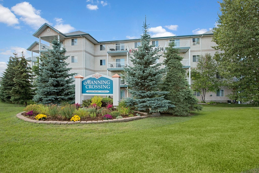 Apartment complex with beige exterior and balconies on four floors. Sign reads "Manning Crossing Apartment Homes" surrounded by manicured garden and trees.