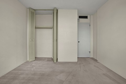 Empty bedroom here at Marquis Towers Apartment Homes with beige walls and carpet. Open closet with light green louvered doors on the left. Closed white door with vent above on the right. Minimalist, calm atmosphere.