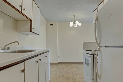 Small, minimalist kitchen here at Marquis Towers Apartment Homes with white cabinets, a stainless steel sink, white appliances including a fridge and stove, and a ceiling light fixture.