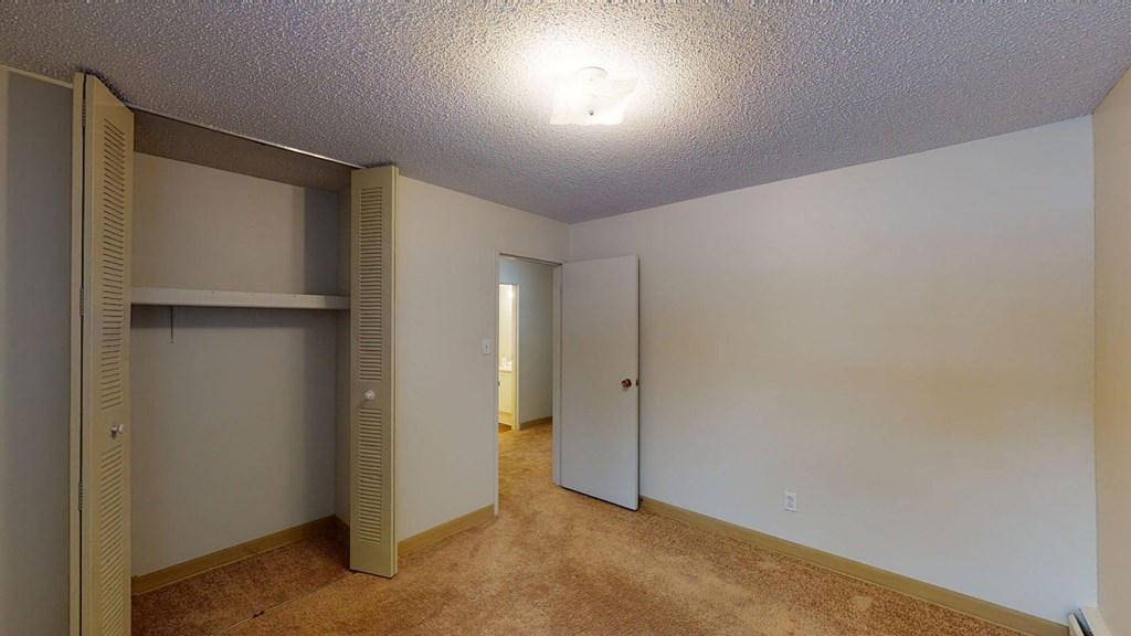 Empty bedroom here at Park Place South Apartment Homes with beige walls and carpeted floor. Open closet with louvered doors on the left. Doorway to a bathroom at the center. Soft overhead light.