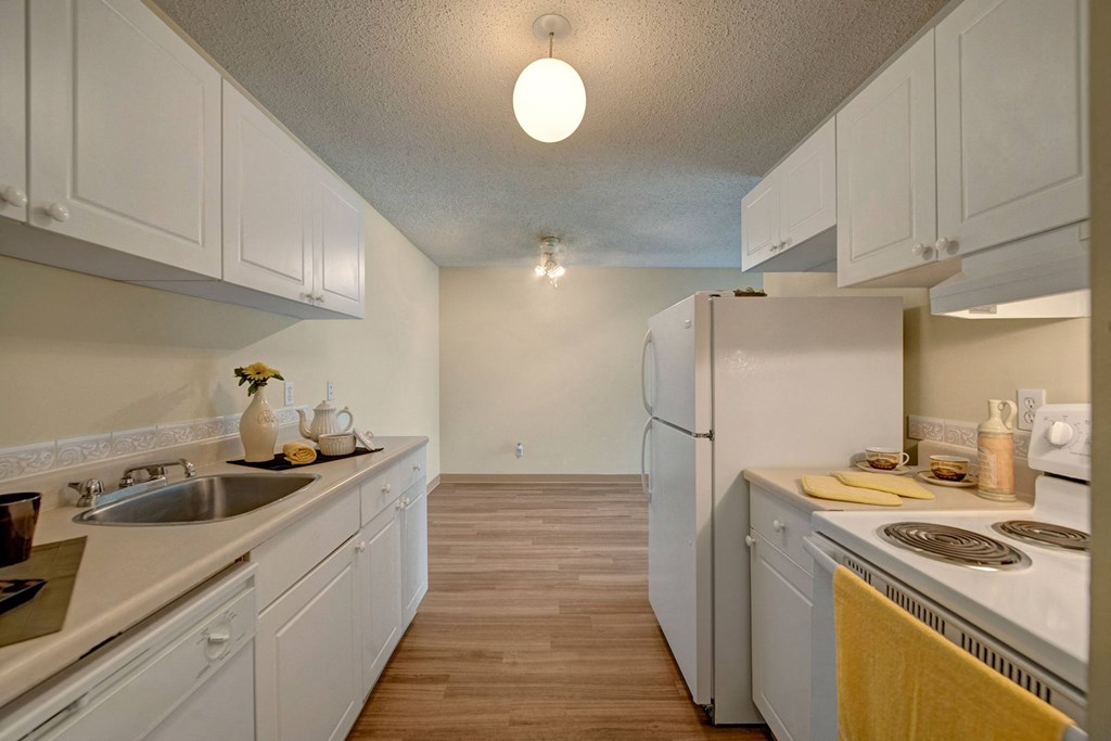 A bright, narrow kitchen here at Park Place South Apartment Homes with white cabinets, a white refrigerator, and stove. A yellow towel hangs near the stove, conveying a clean, minimalist feel.