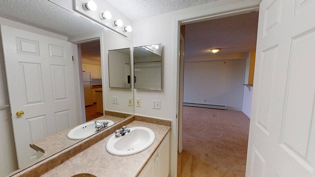 Bathroom here at The Pointe at Applewood with beige countertop, sink, and mirror. Bright lights above. Open door reveals a kitchen with white cabinets and an adjacent room. Cozy and inviting.