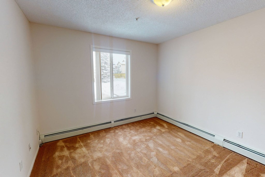Empty bedroom here at The Pointe at Applewood with beige walls and a brown carpet. A window on the left shows a snowy outdoor scene. Soft lighting creates a calm, inviting atmosphere.
