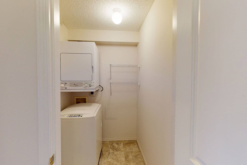 Compact laundry room here at The Pointe at Applewood with a stackable washer and dryer, beige walls, and a bright overhead light. Two wire shelves are mounted on the right wall.