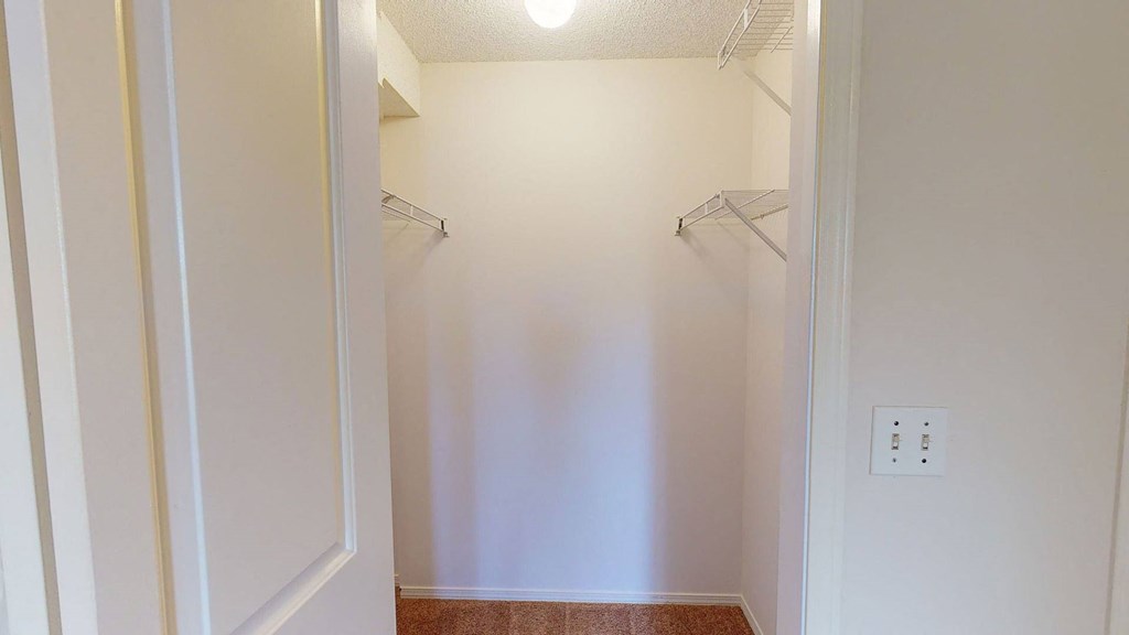 Empty closet here at The Pointe at Applewood with beige walls and a beige carpeted floor, illuminated by a ceiling light. Metal racks are mounted on the left and right sides.