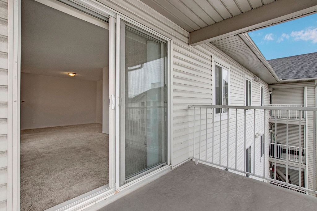 View of a small balcony here at The Pointe at Applewood with white railing attached to a white building, leading to an empty room with beige carpet and a ceiling light.
