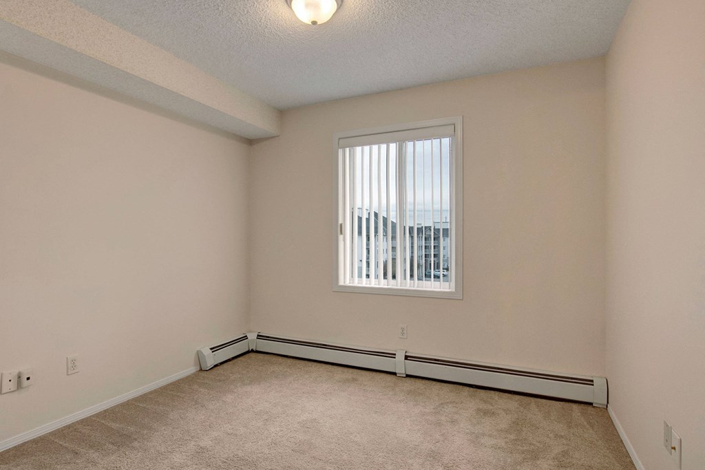 Empty room here at The Pointe at Applewood with beige walls and carpet, brightened by a single window with vertical blinds. The room has a ceiling light and baseboard heaters.