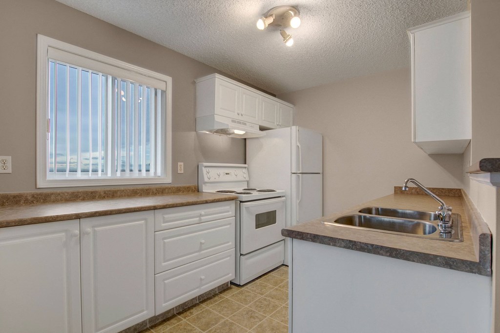 Compact kitchen here at The Pointe at Applewood with beige walls, featuring white cabinets, stove, and refrigerator. Double sink on a brown countertop. Bright window with blinds.
