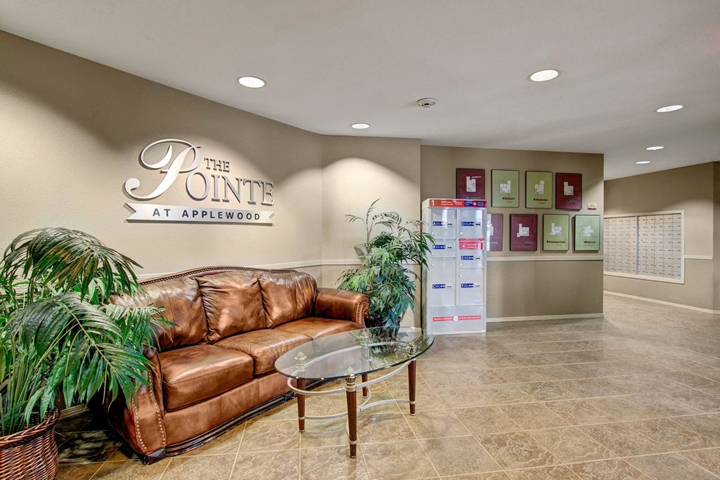 Cozy lobby with a brown leather couch, glass coffee table, and potted plants. Wall sign reads "The Pointe at Applewood." Mailboxes in background.