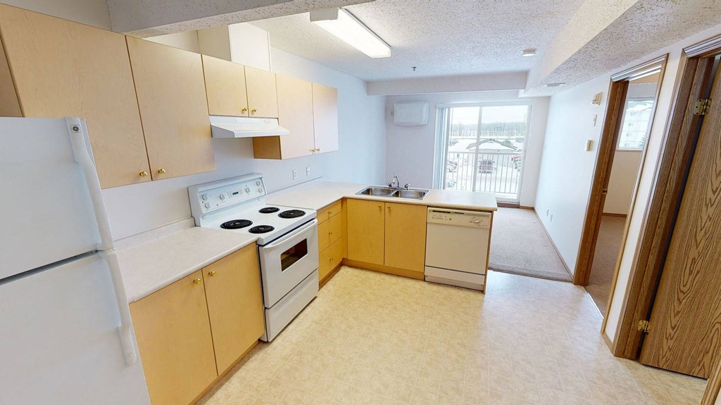Bright open kitchen here at Quail Landing Apartment Homes with light wood cabinets, white appliances, and a double sink. Large window view, carpeted living space. Clean, modern feel.