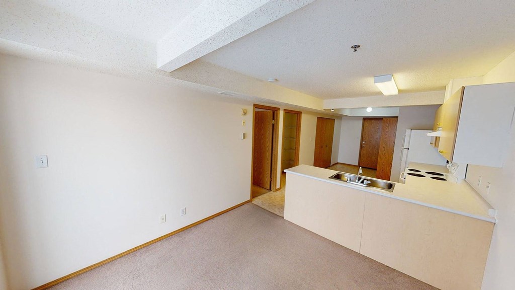 A clean, empty room here at Quail Landing Apartment Homes with beige carpet and walls, featuring a small kitchen area with white cabinets, a stove, and a double sink. Two wooden doors lead to other rooms.