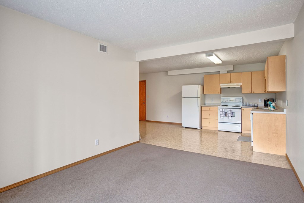 Empty living area here at Quail Landing Apartment Homes with beige carpet and a simple kitchen. The kitchen features light wooden cabinets, a white fridge, stove, and minimal decor.