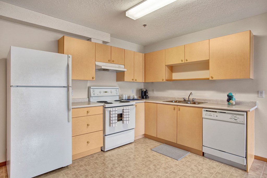 Modern kitchen here at Quail Landing Apartment Homes with light wood cabinets, white refrigerator, stove, and dishwasher. Coffee maker on counter, bright and tidy ambiance.