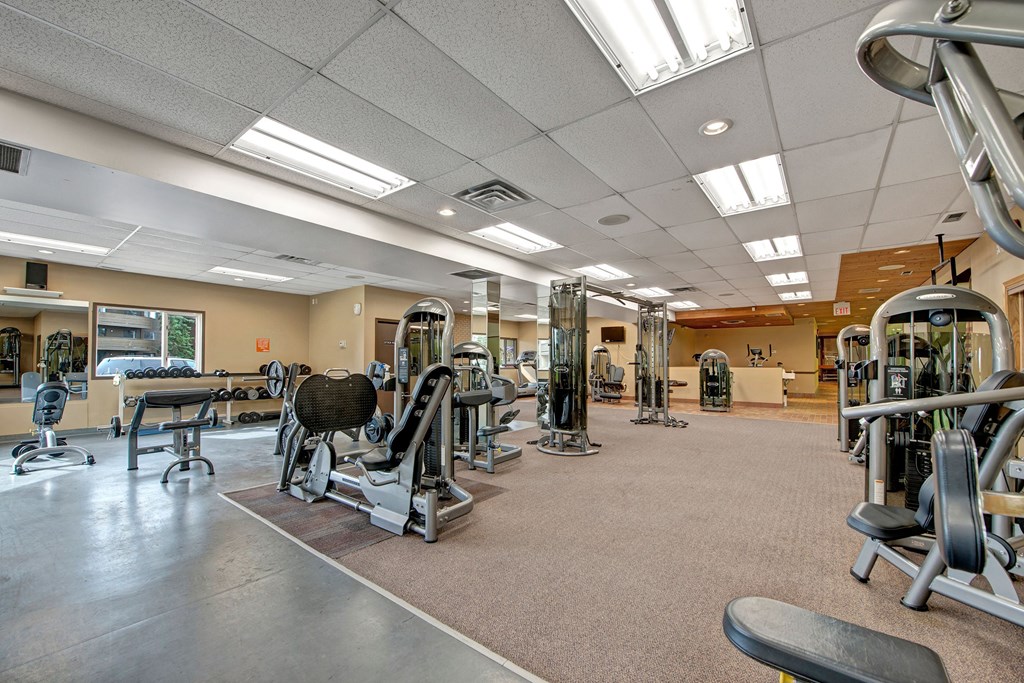 Bright gym interior here at Ranchlands Apartment Homes with various exercise machines, mirrored walls, and light gray flooring. The atmosphere is clean and organized, inviting fitness activity.