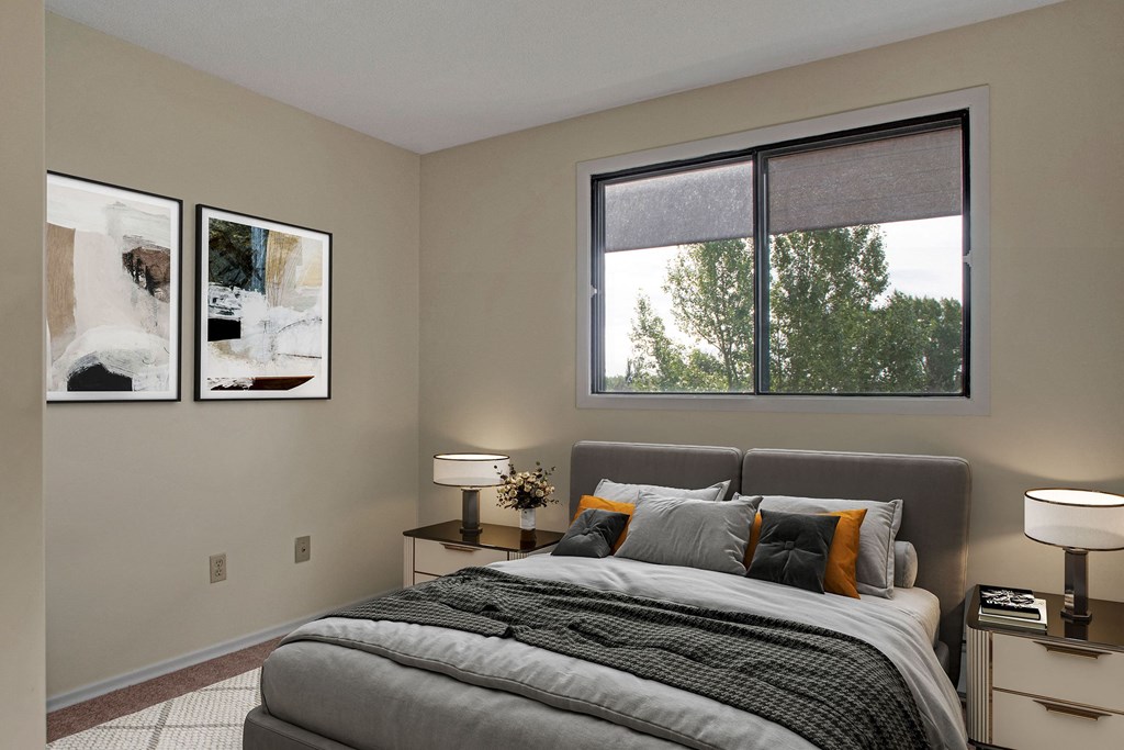 Modern bedroom here at Ranchlands Apartment Homes with a soft gray bed, orange accent pillows, and abstract art on beige walls. Large window reveals lush green trees outside. Cozy and serene atmosphere.