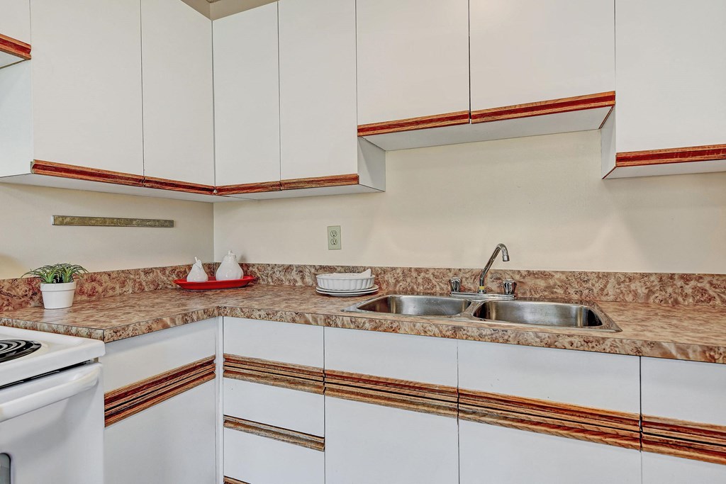 A bright kitchen here at Ranchlands Apartment Homes with white cabinets, wood accents, and a marbled countertop. Double sink with a chrome faucet. Items include a plant and white teapots on a red tray, creating a cozy atmosphere.