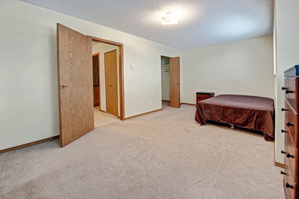 A bedroom with a large closet and overhead lighting Spacious bedroom here at Ridgeview Village Apartment Homes with beige carpet, light walls, and wood doors. A bed with a brown cover is against the wall, and a dresser is on the right. Cozy and minimalist.