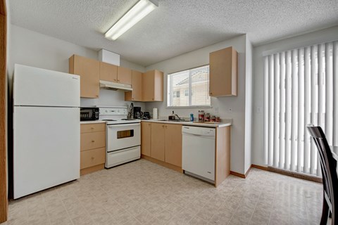Compact kitchen here at Ridgeview Village Apartment Homes with white appliances, beige cabinets, and tile flooring. Bright light from a window and vertical blinds adds a cheerful ambiance.