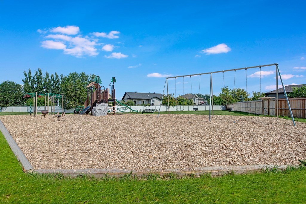 Playground here at Sol Terrace Apartment Homes with swings and climbing set, surrounded by wood chips and grass. Houses and trees in the background under a bright blue sky.