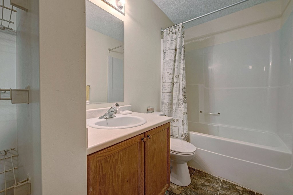 Small bathroom here at Sol Terrace Apartment Homes with wood cabinet, sink, and toilet. A white shower curtain with decorative patterns hangs over a bathtub. Light tones create a clean, simple look.