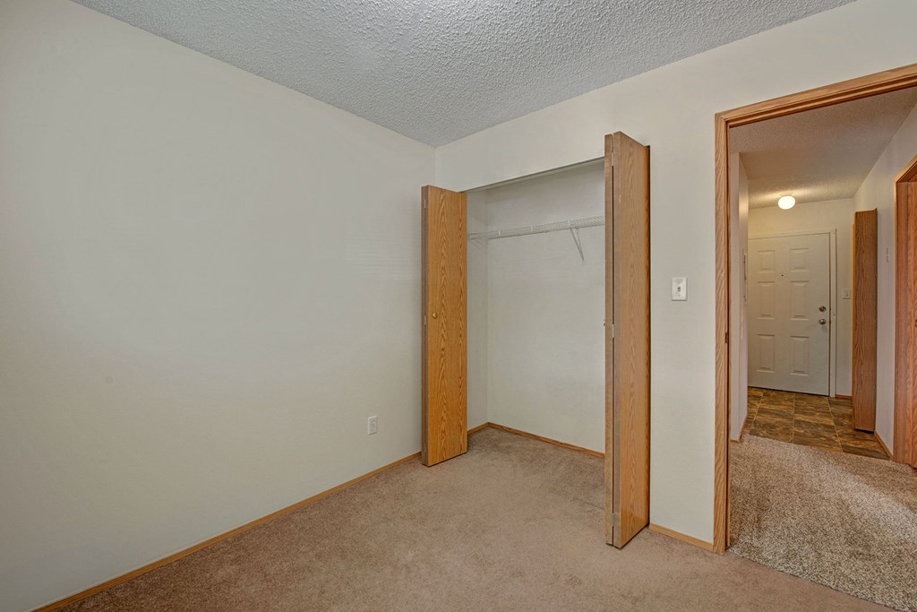 Empty room here at Sol Terrace Apartment Homes with beige carpet, open wooden closet doors revealing a shelf, and an entrance leading to a lit hallway with a closed white door. Simple and neutral atmosphere.