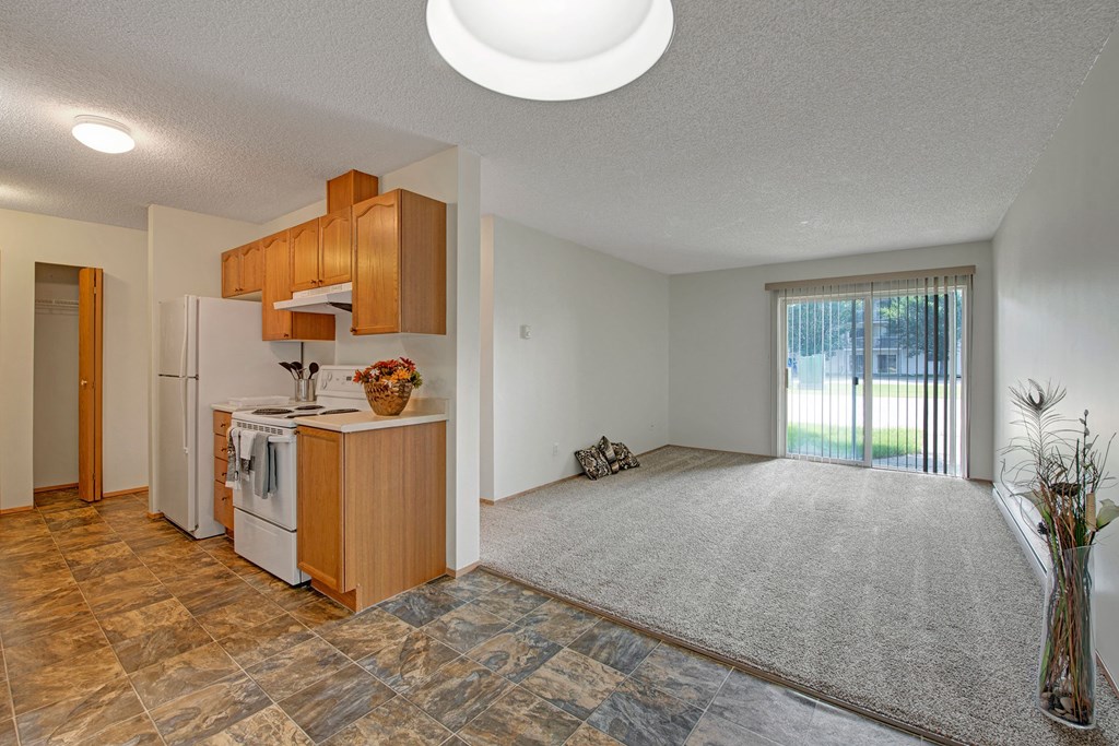 Modern open-plan apartment here at Sol Terrace Apartment Homes with brown tile kitchen, oak cabinets, and carpeted living area. Sliding glass doors lead to a bright, grassy yard.