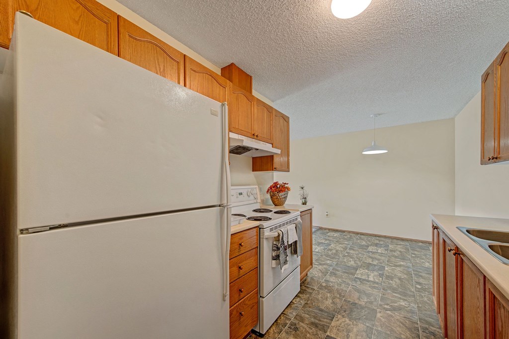 Simple kitchen here at Sol Terrace Apartment Homes with wooden cabinets and gray-tiled floor. Features a white fridge, stove, and a countertop with a basket of flowers, under warm lighting.