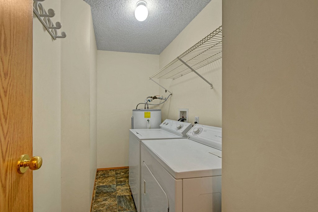 Laundry room here at Sol Terrace Apartment Homes with a washing machine, dryer, and water heater. An overhead shelf is mounted on the right wall. The room has beige walls and tiled floor.