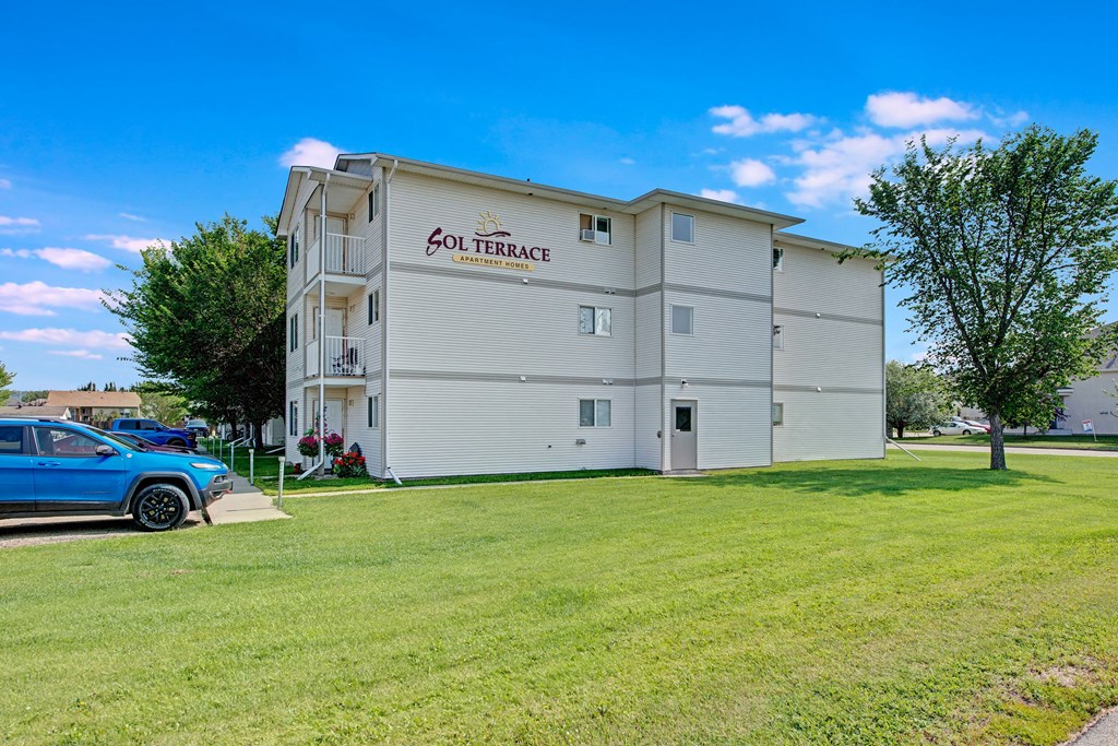 Four-story beige apartment building, labeled "Sol Terrace," with well-maintained lawn, trees, and a parked blue SUV under a bright blue sky.
