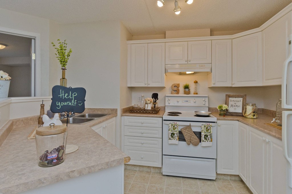 Cozy kitchen here at Somerset Pointe Apartment Homes with white cabinets, a stove, and decor items like plants and framed art. A "Help yourself" sign sits on a counter with a candy jar.