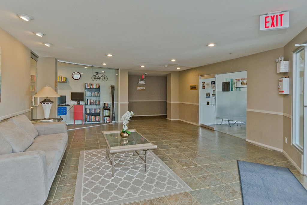Cozy lobby here at Somerset Pointe Apartment Homes with a beige sofa, glass coffee table on a patterned rug, and bookshelves. Bright lighting creates a welcoming atmosphere; exit sign visible.