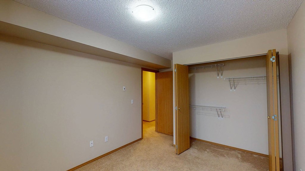 Empty beige room here at Sonora Apartment Homes with a textured ceiling and a closed door to the hallway. An open closet has shelves and wooden bifold doors, under warm lighting.