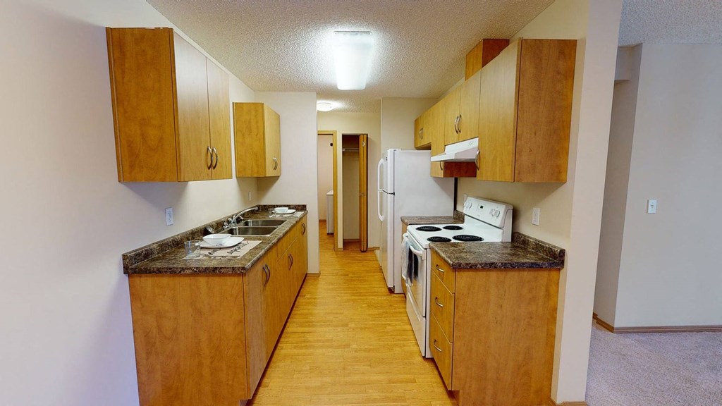 Narrow kitchen here at Sonora Apartment Homes with wooden cabinets, marble countertops, and white appliances. Bright lighting and a cozy, organized atmosphere.