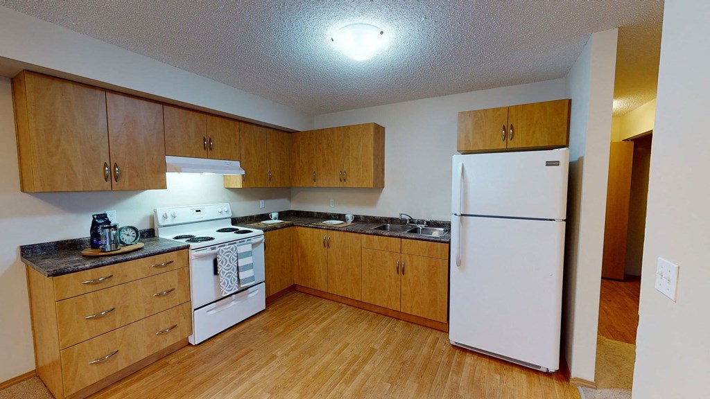 Compact kitchen here at Sonora Apartment Homes with light wood cabinets, white appliances, and brown countertops. Includes a stove, fridge, and double sink, creating a cozy atmosphere.