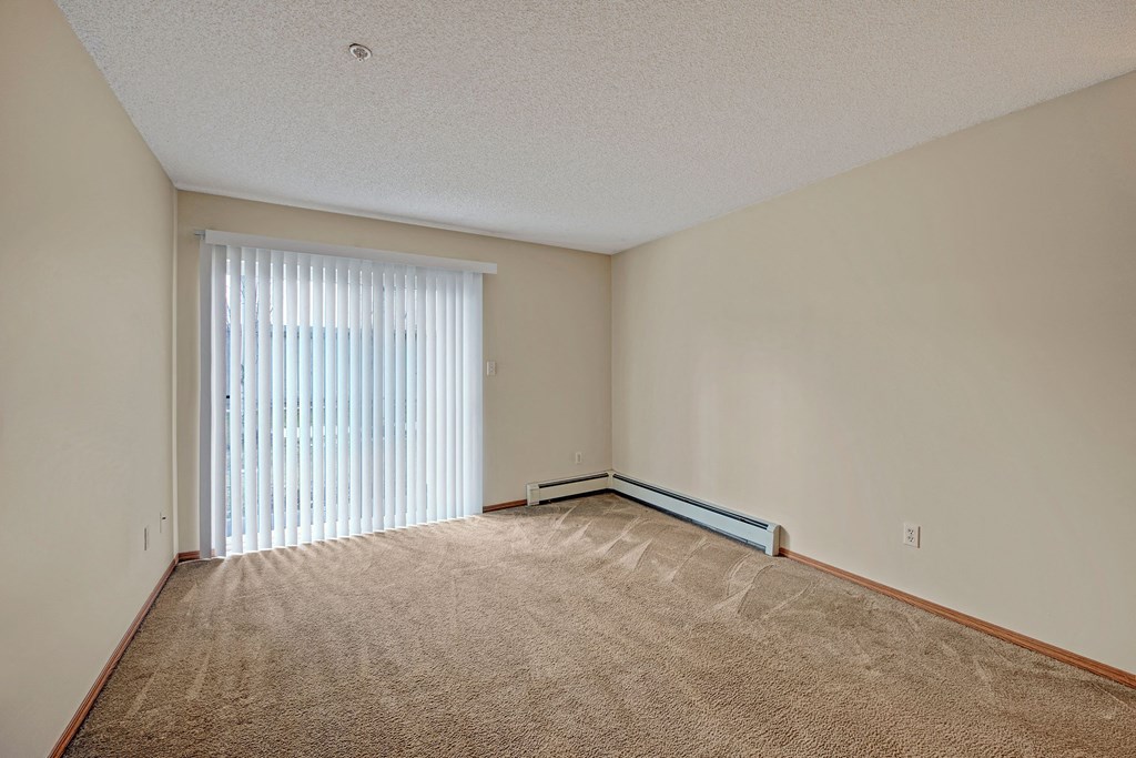 Empty room here at Sonora Apartment Homes with beige walls and carpet, featuring a large window covered by vertical blinds. Sunlight softly illuminates the space, creating a calm ambiance.