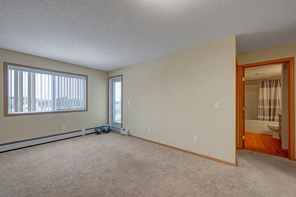 Empty carpeted room here at Sonora Apartment Homes with beige walls, large window with vertical blinds on the left, and a door leading to a bathroom on the right. Neutral, calm ambiance.