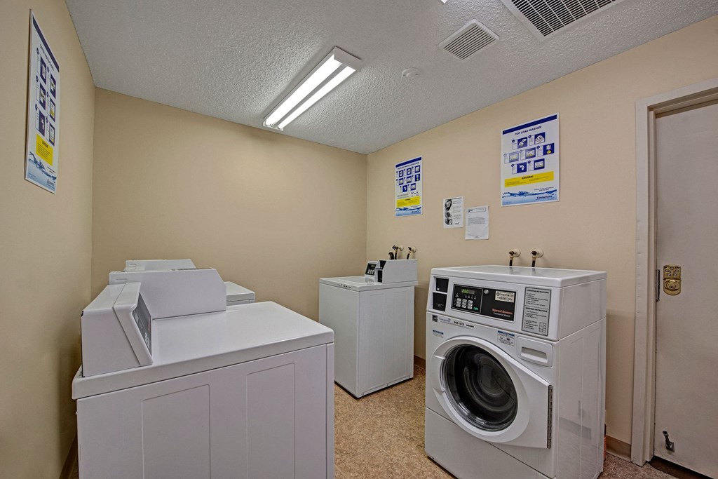 Laundry room here at Sterling Place Apartment Homes with two top-loading washers and a front-loading dryer. Beige walls, fluorescent lighting, and instructional posters hang on the walls. Clean and functional atmosphere.