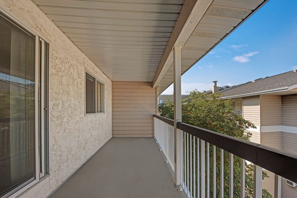 Covered balcony here at Sterling Place Apartment Homes with beige stucco walls, sliding glass door, and black railing. Overlooks a tree and neighboring beige building under a clear blue sky.