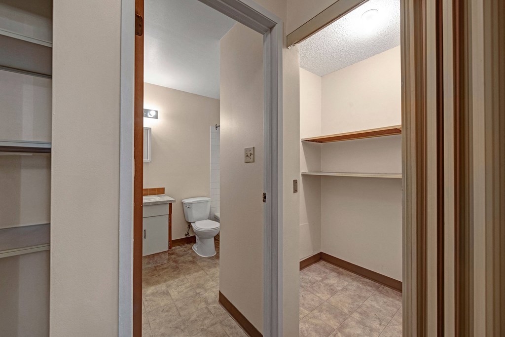 A narrow hallway here at Sterling Place Apartment Homes leading to a bathroom with a toilet, white tiles, and a small vanity with a light above, and a closet with empty shelves on the right.