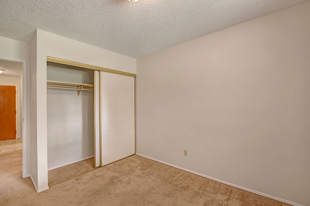 Empty room here at Sterling Place Apartment Homes with beige carpet and light beige walls. A small closet with sliding doors is on the left, next to an open door leading to a hallway.