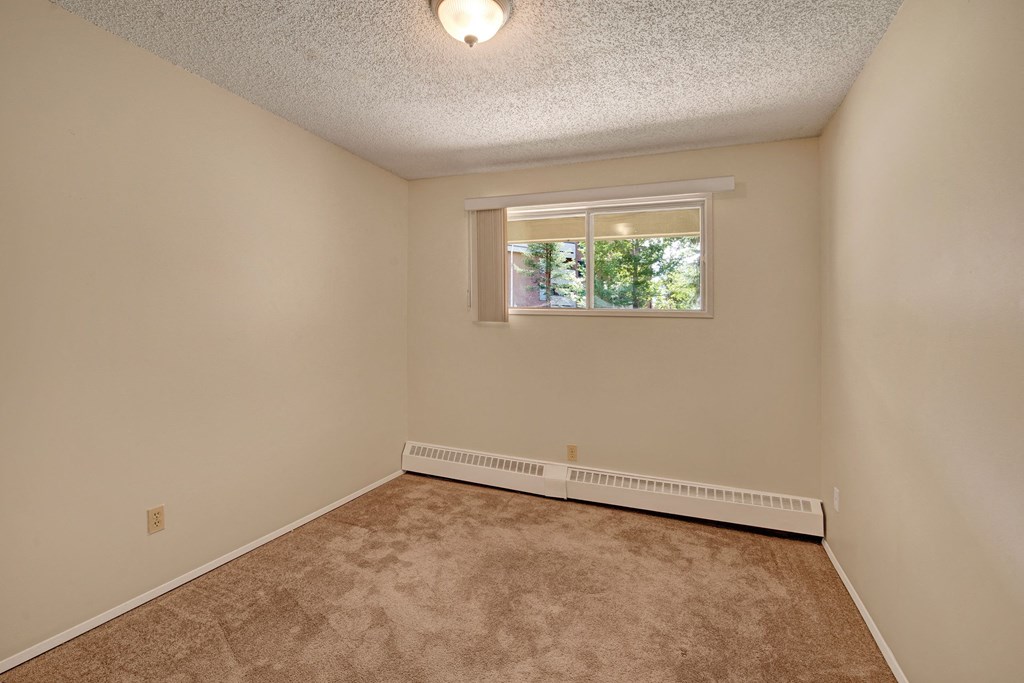 Empty room here at Sterling Place Apartment Homes with beige walls and carpet, a window with blinds partially open, and a ceiling light. Natural light creates a calm, inviting atmosphere.